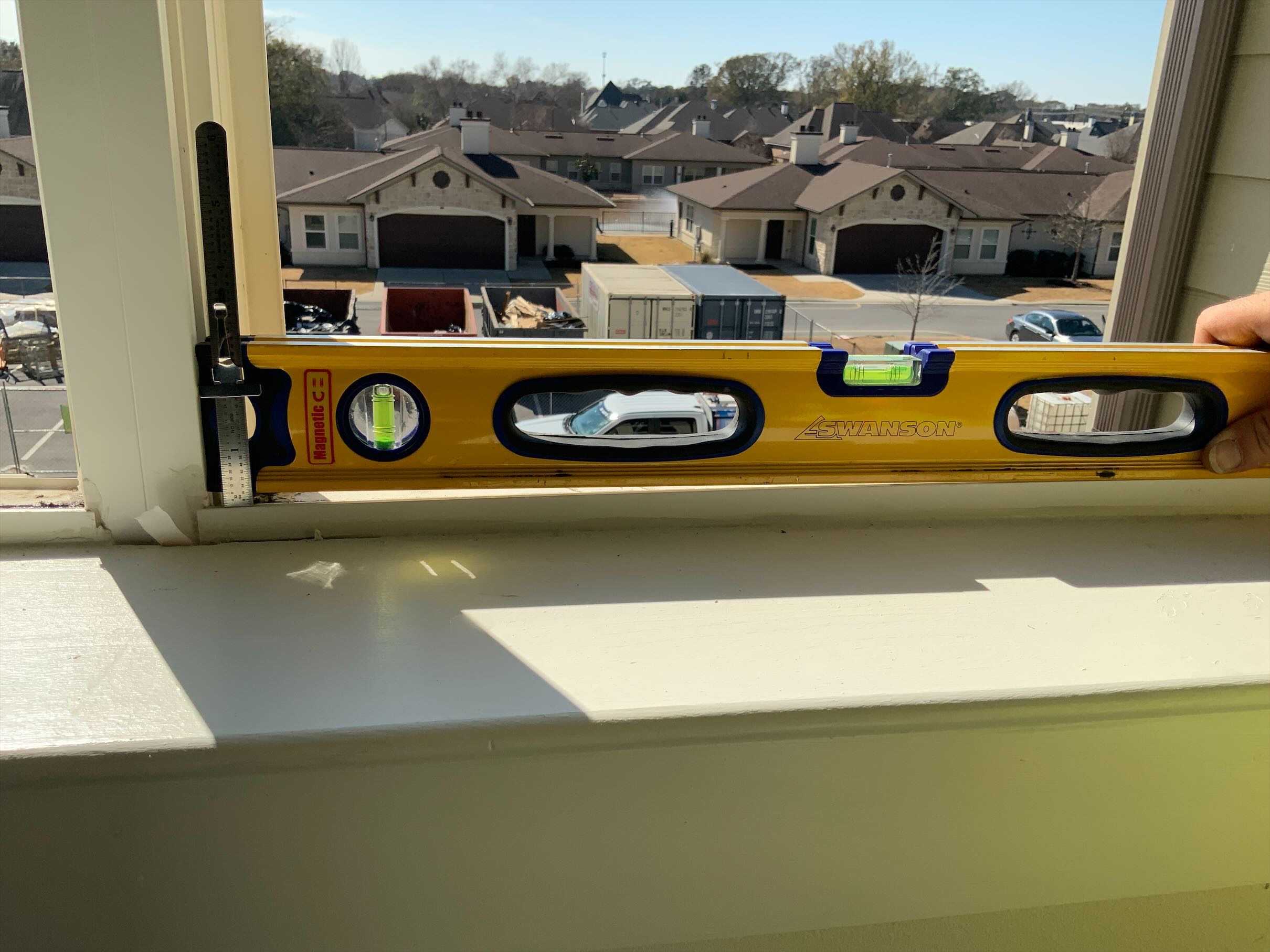 A person uses a yellow level tool to align a window sill. The scene overlooks a suburban neighborhood with several rooftops in sunlight.