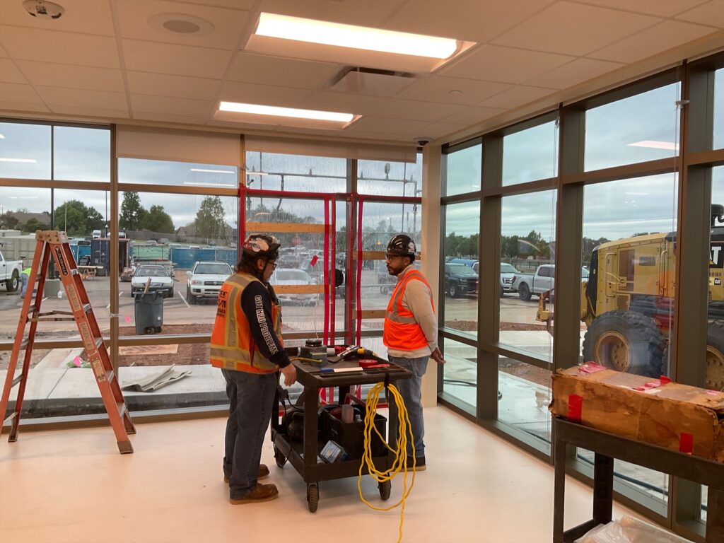 Two construction workers in safety vests and helmets stand indoors near large windows and equipment. Outside, a construction site and vehicles are visible.