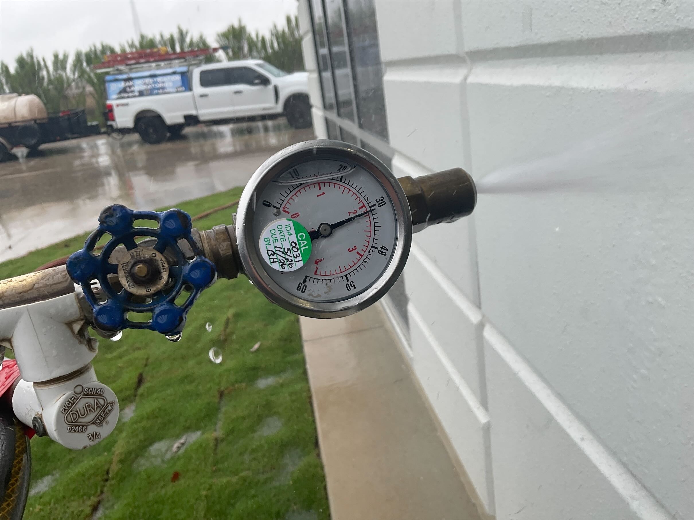 A pressure gauge with water spraying from a pipe, set against a rainy backdrop. A white truck and green grass are visible, conveying a utility area.