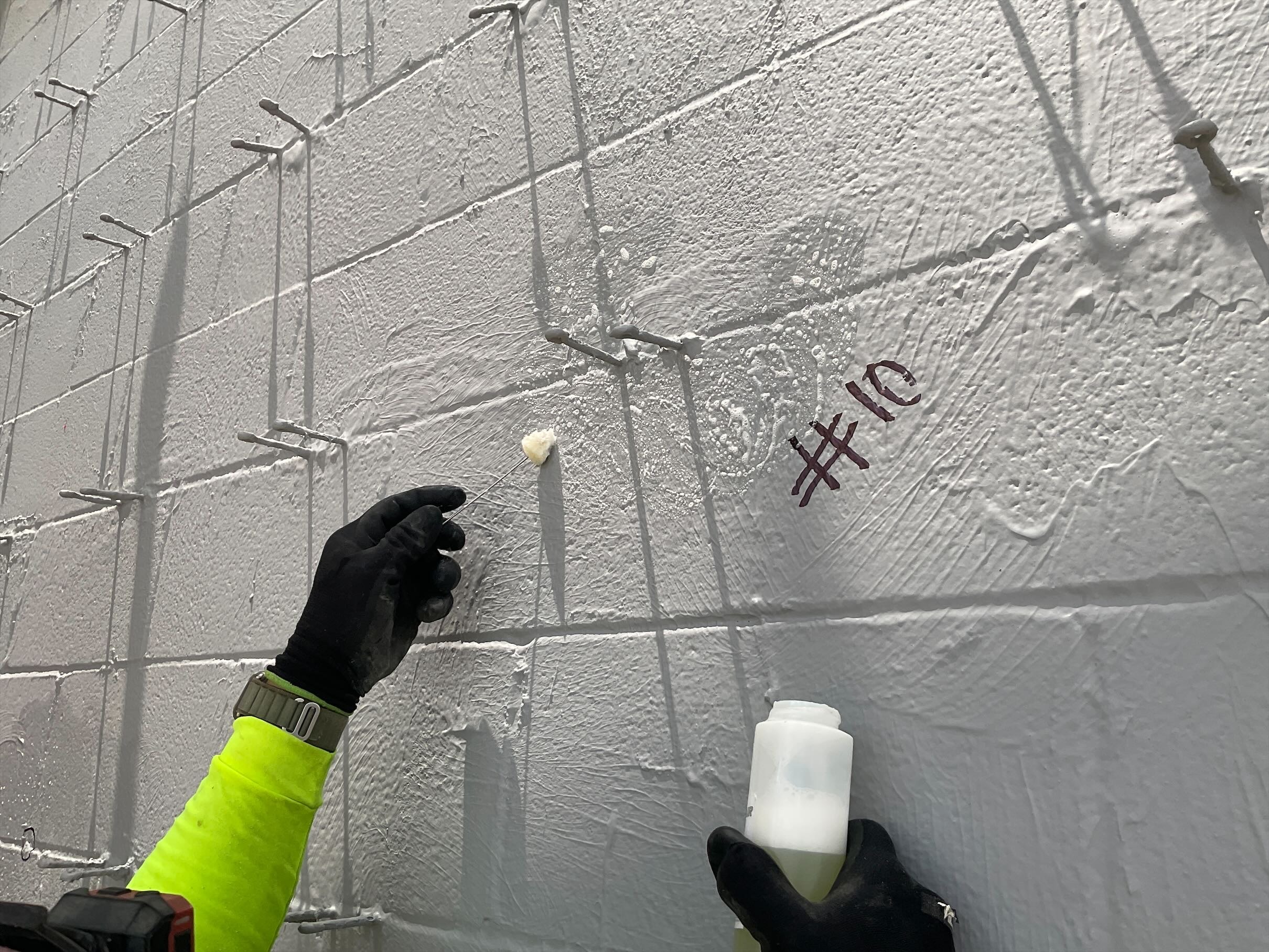 Gloved hands applying substance to a white concrete wall with protruding metal rods. The number "#10" is written on the wall.