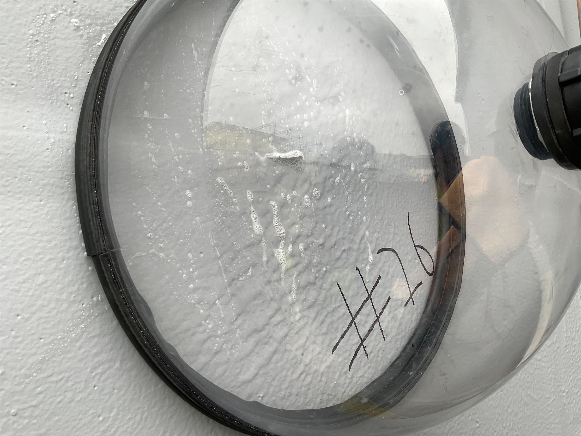 A round, clear dome with a black rim showing water droplets on the surface, reflecting a hand holding a device. The number "#26" is written on it.