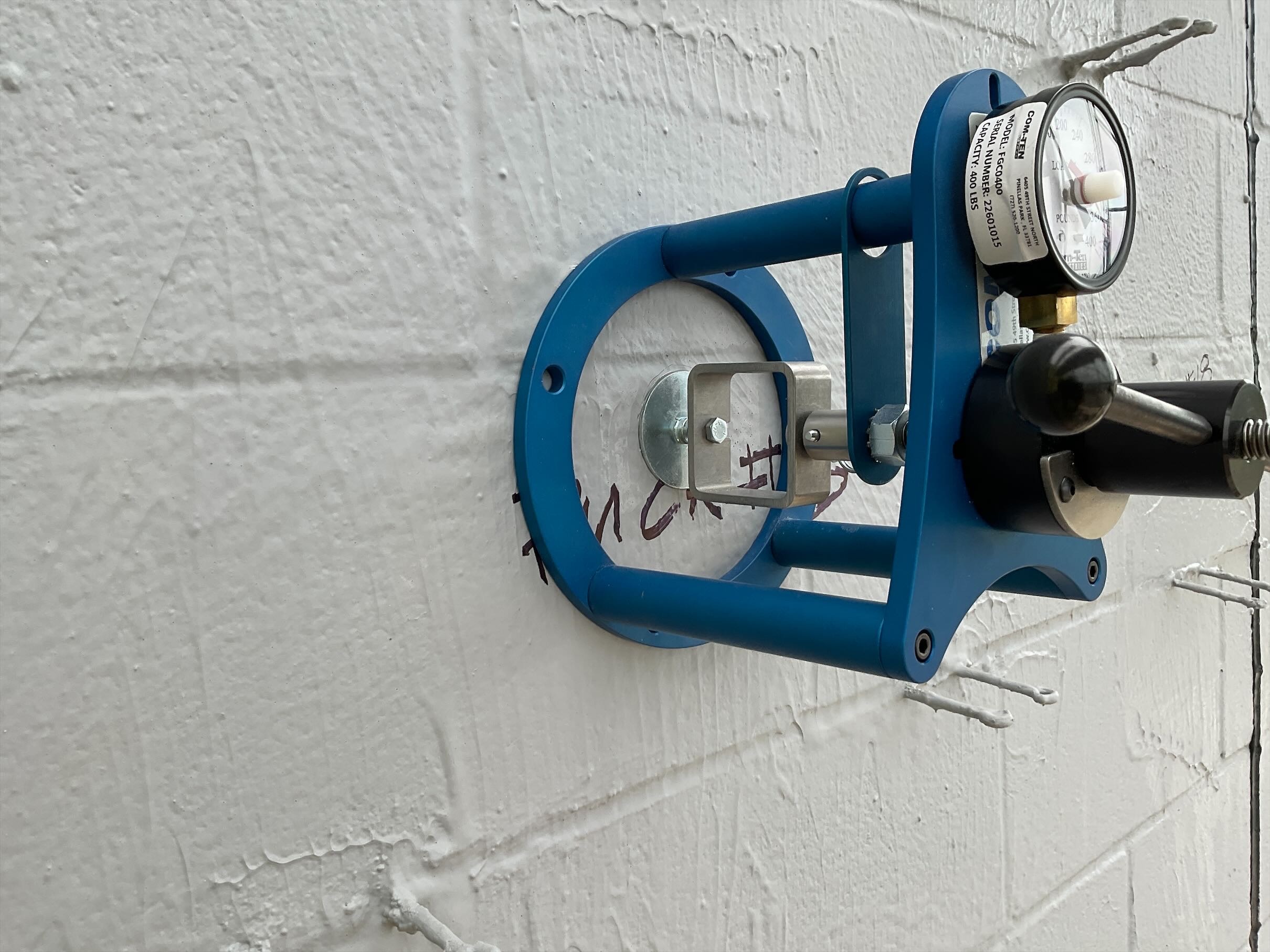 A blue metal bracket is mounted on a white brick wall, holding a pressure gauge and valve. The setup appears technical and industrial.