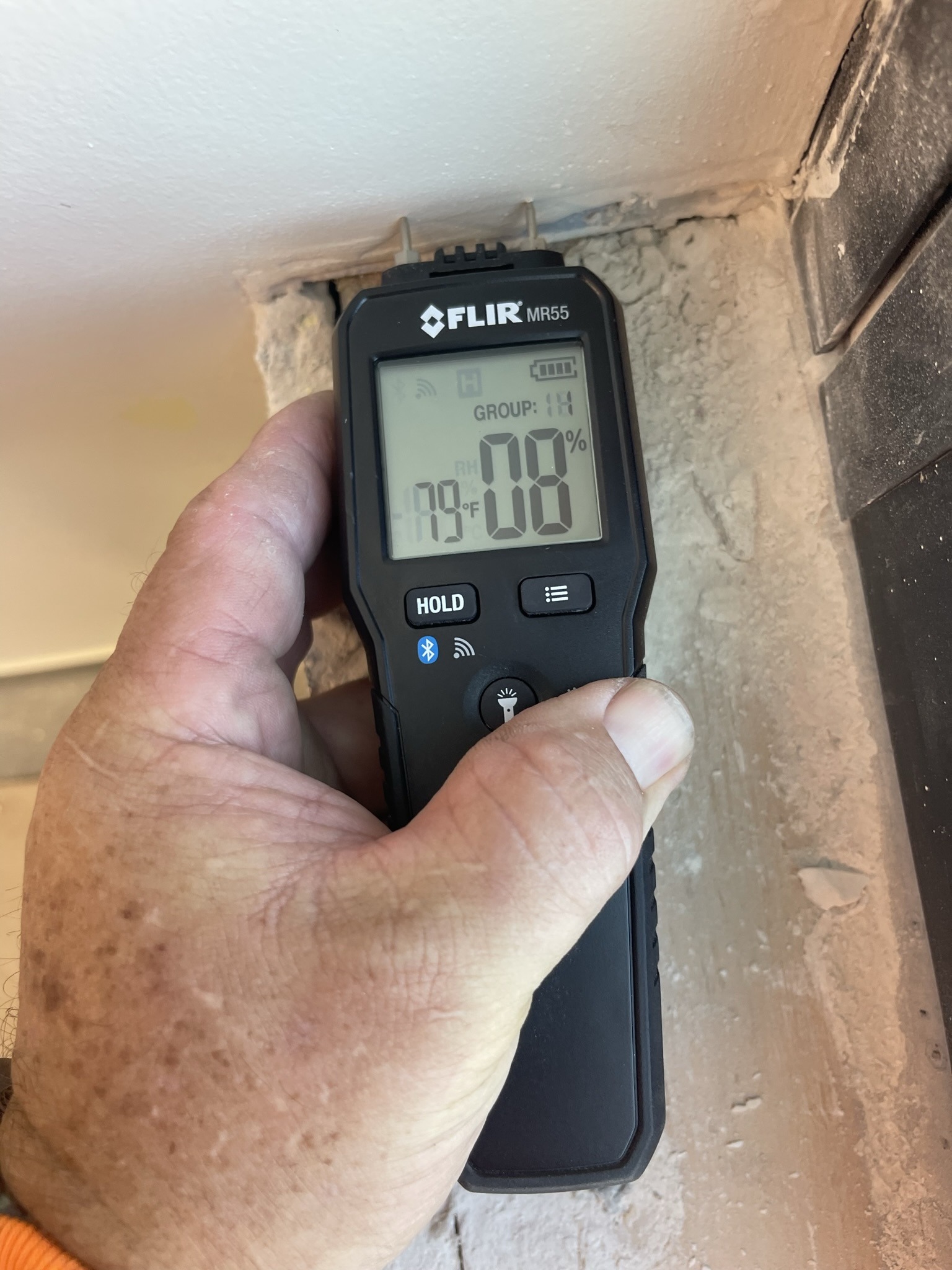A hand holds a digital moisture meter against a textured wall corner. The device displays a moisture level of 8% and a temperature of 79°F.