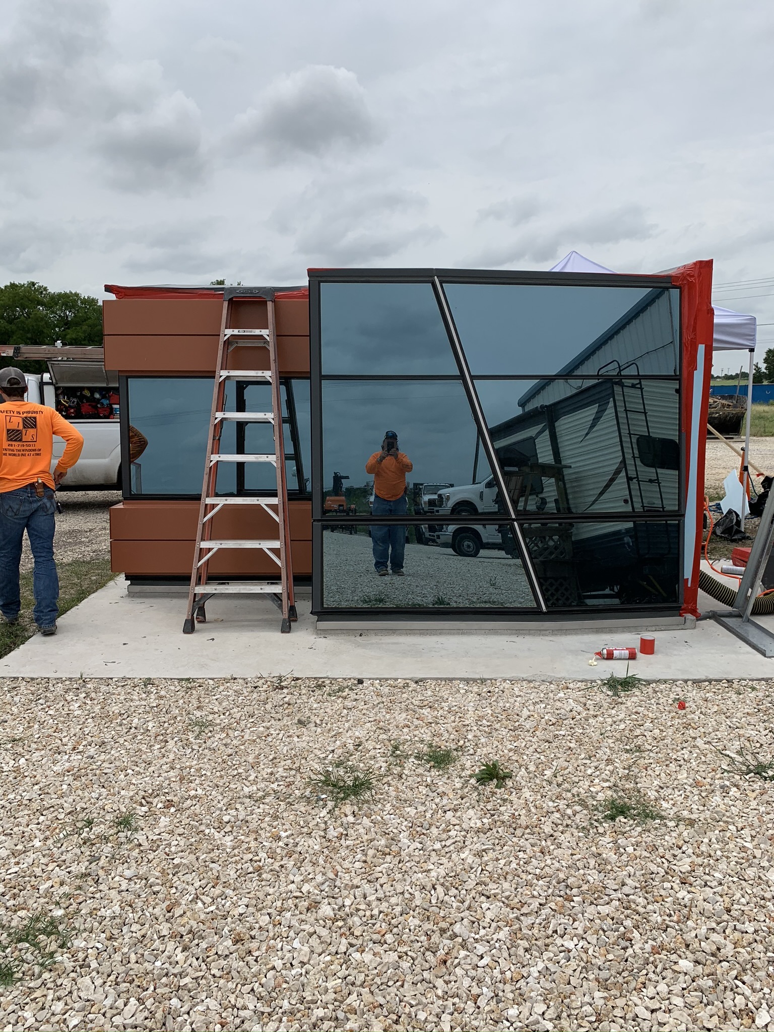 A man in an orange shirt is reflected in large, angular glass panels of a modern building facade. A ladder rests against the structure, and the sky is overcast.