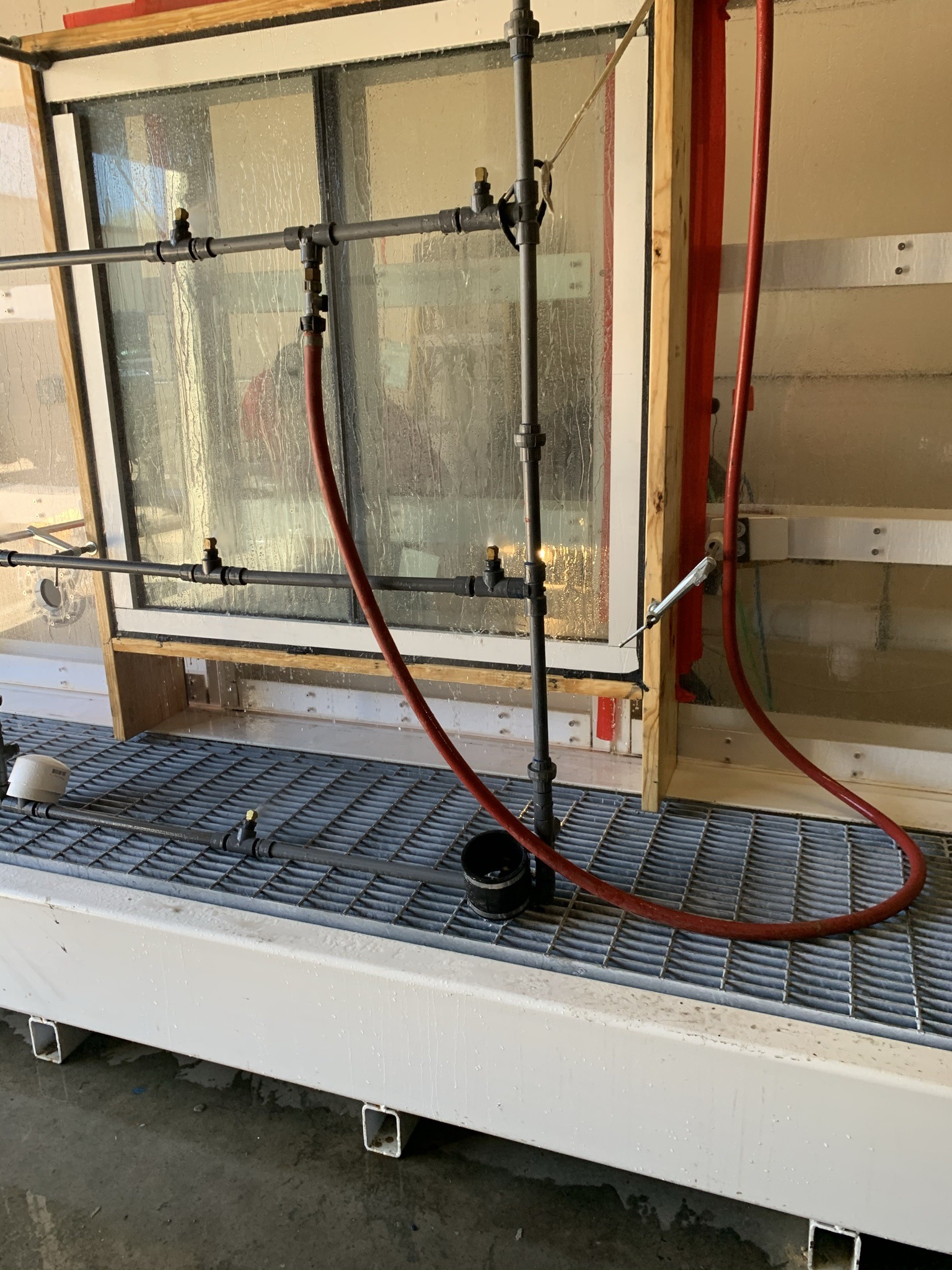 Wet industrial testing setup with water spraying on a large glass panel in a metal frame. Red hoses and black pipes are visible on a grated floor.