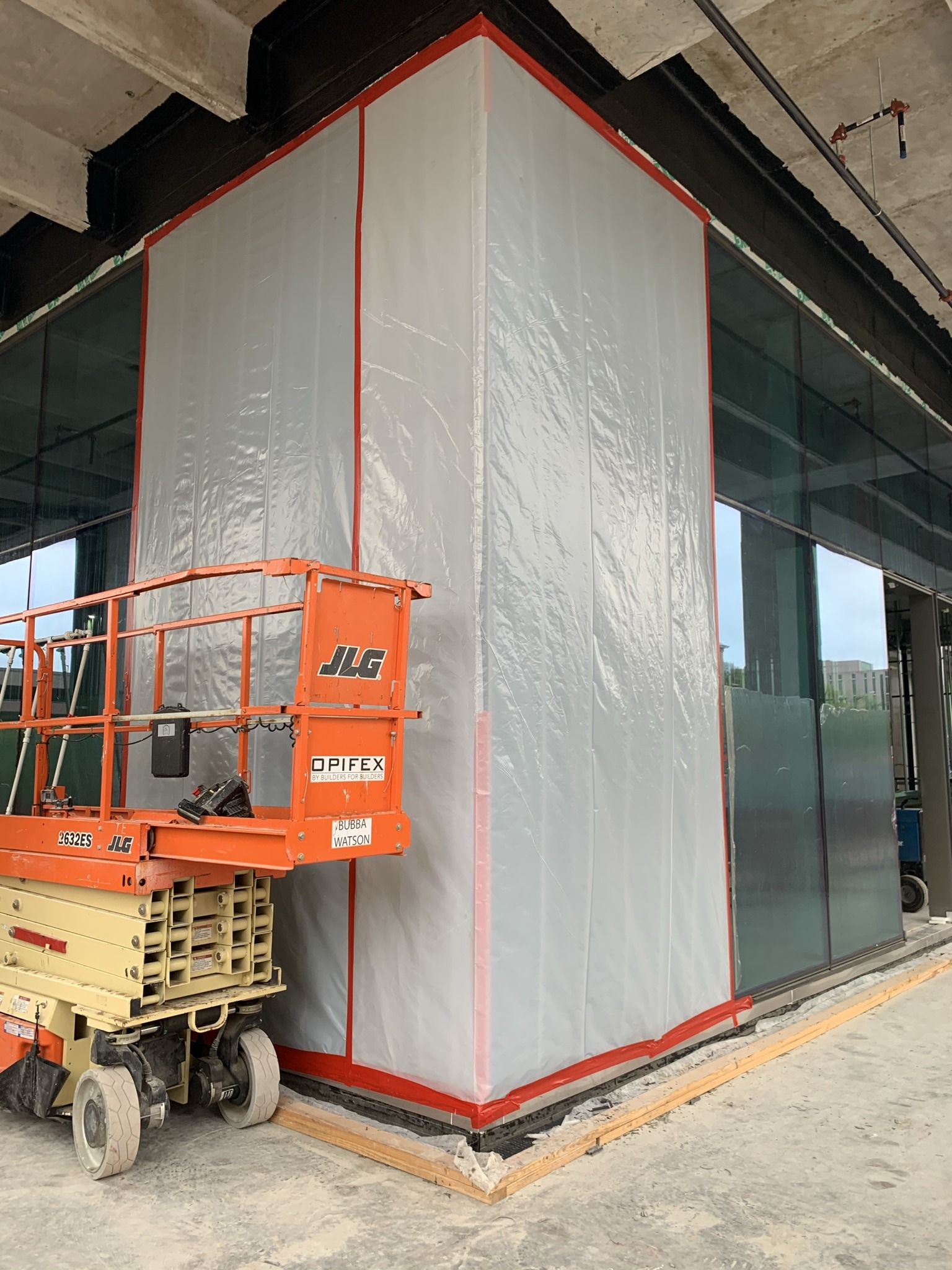 An orange scissor lift is parked beside a corner of a building under construction, covered in plastic sheeting secured with red tape. The scene feels industrial and in-progress.