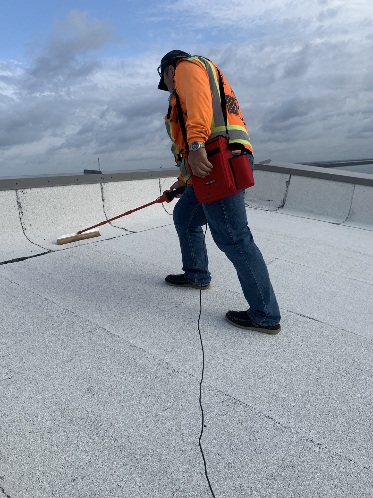 A worker in jeans and an orange safety vest inspects a flat rooftop with a long-handled tool, set against a cloudy sky.